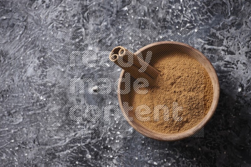 Wooden bowl full of cinnamon powder and a cinnamon stick on a textured black background