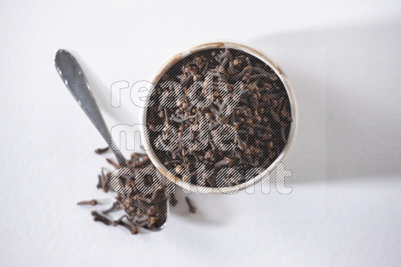 A beige ceramic bowl full of cloves and a metal spoon next to it on a white flooring