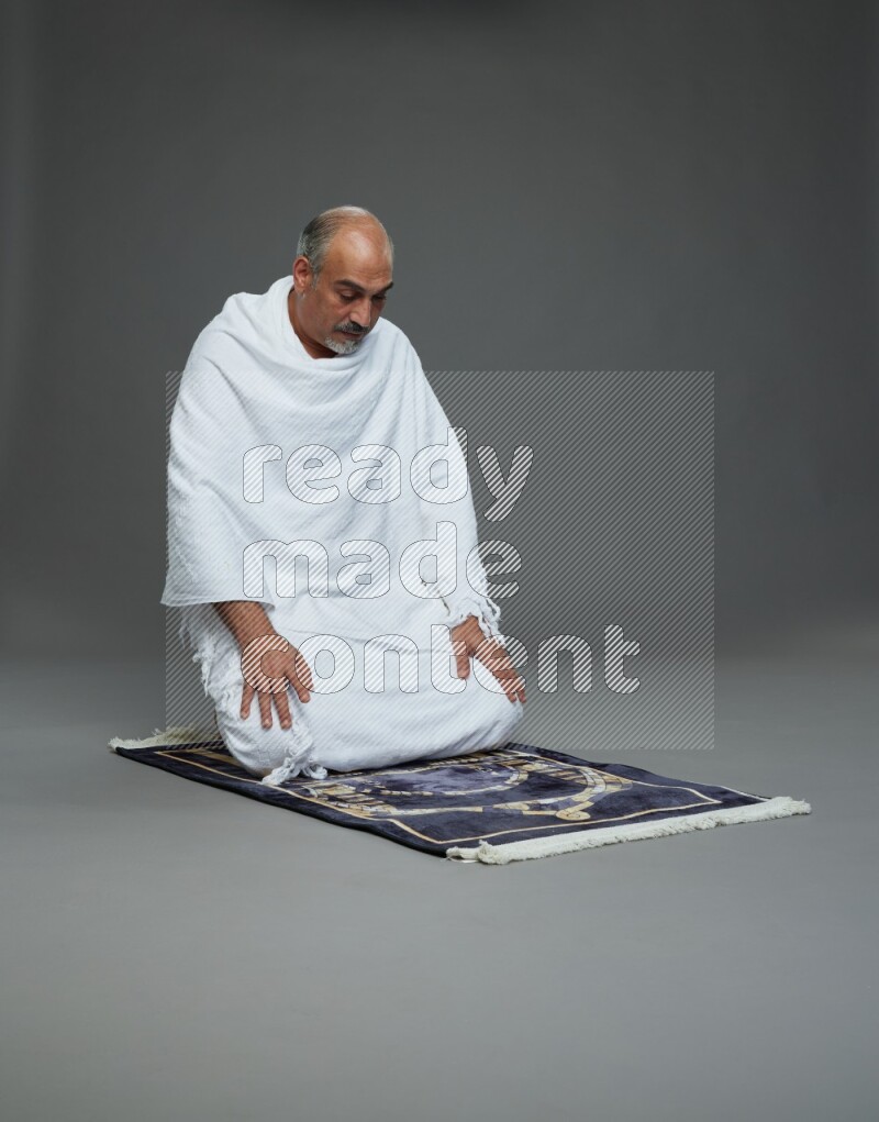 A man wearing Ehram sitting on prayer mat on gray background
