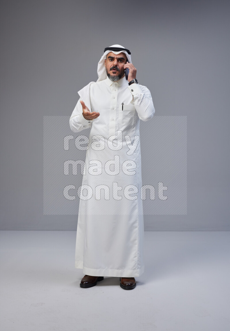 Saudi man Wearing Thob and white Shomag standing talking on phone on Gray background
