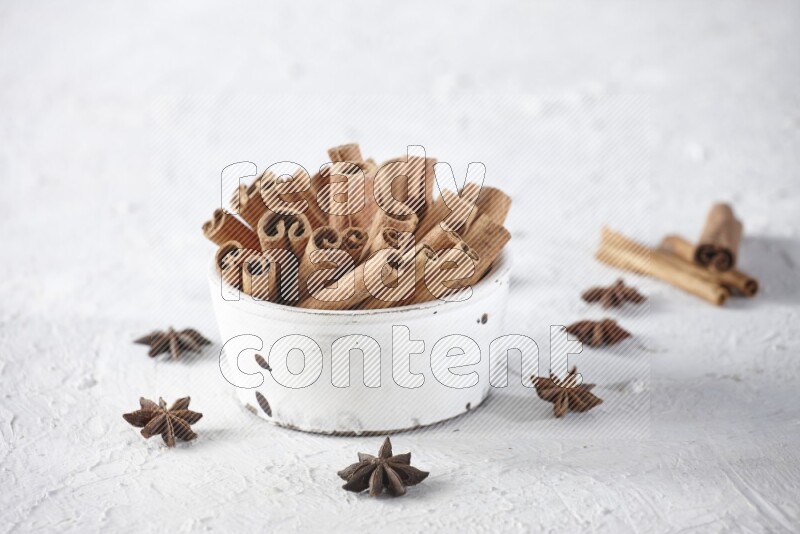 White bowl full of cinnamon sticks surrounded by star anis on a textured white background in different angles