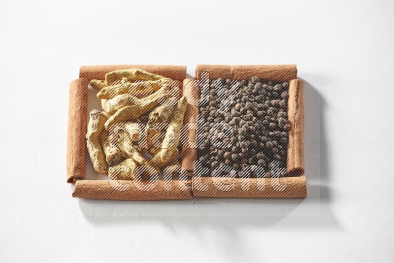 مربعان من أعواد القرفة ممتلئان بحبوب الفلفل الأسود وأصابع الكركم علي خلفية بيضاء