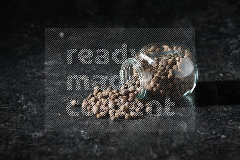 A flipped glass spice jar full of allspice whole balls and the balls spilled out of it on a textured black flooring