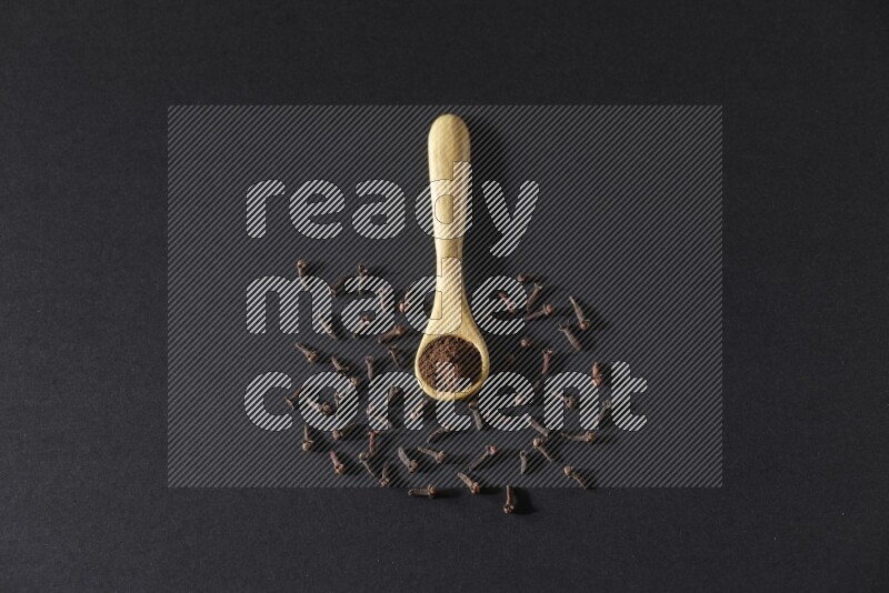 A wooden spoon full of cloves powder with spreaded whole cloves on a black flooring