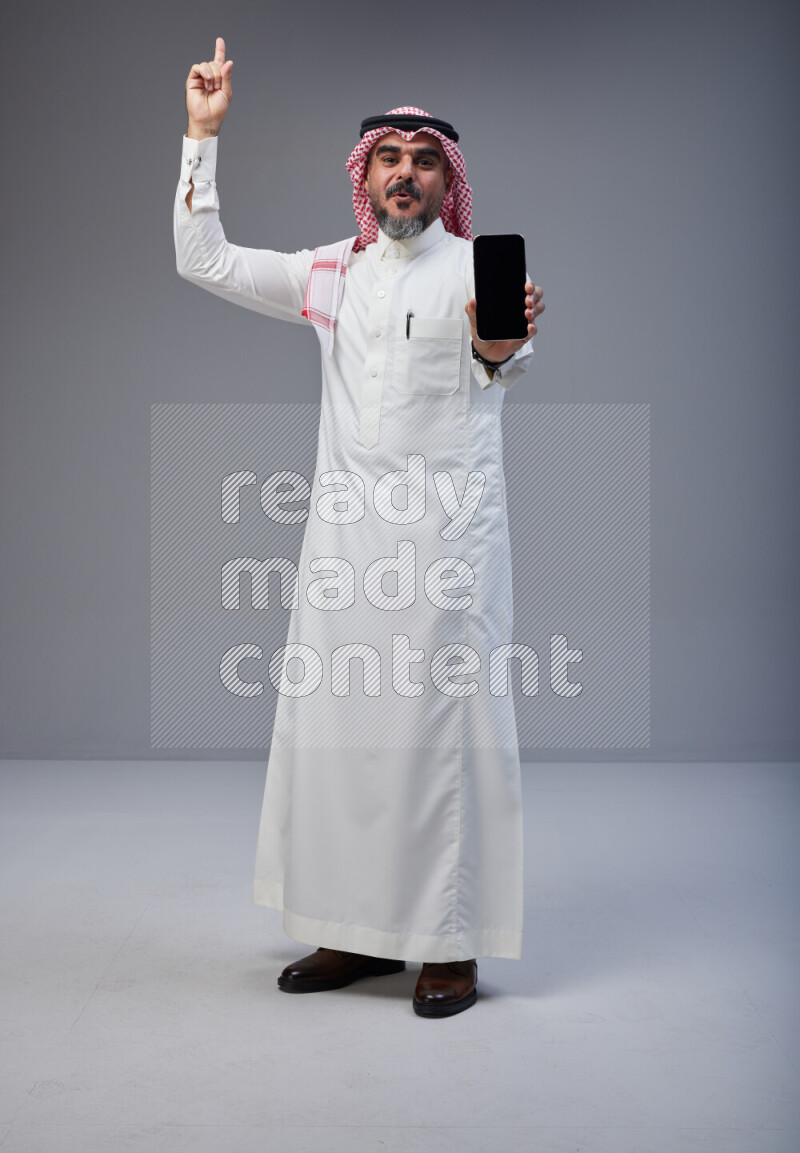 Saudi man Wearing Thob and red Shomag standing showing phone to camera on Gray background