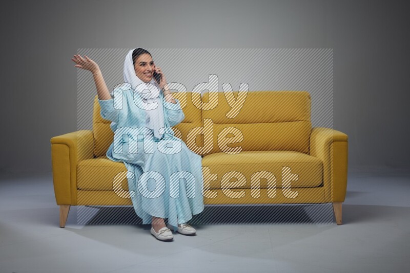 A Saudi woman wearing a light blue Abaya and a white head scarf sitting on a yellow sofa and talking in the phone while making different poses eye level on a grey background
