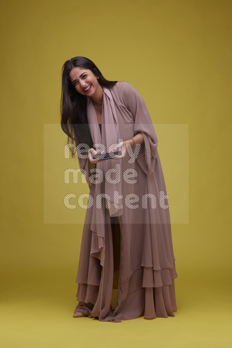 A woman Playing a Game on her smartphone on a Yellow Background wearing Brown Abaya