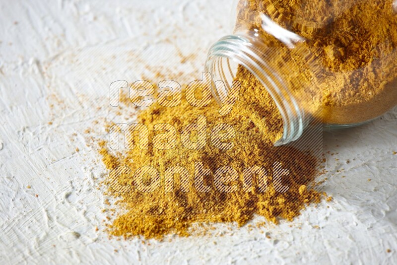 A flipped glass spice jar full of turmeric powder and powder fell out it on textured white flooring