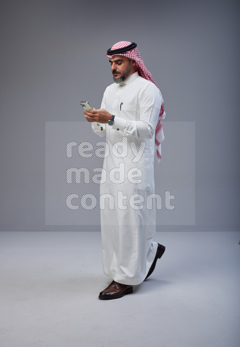 Saudi man Wearing Thob and red Shomag standing texting on phone on Gray background