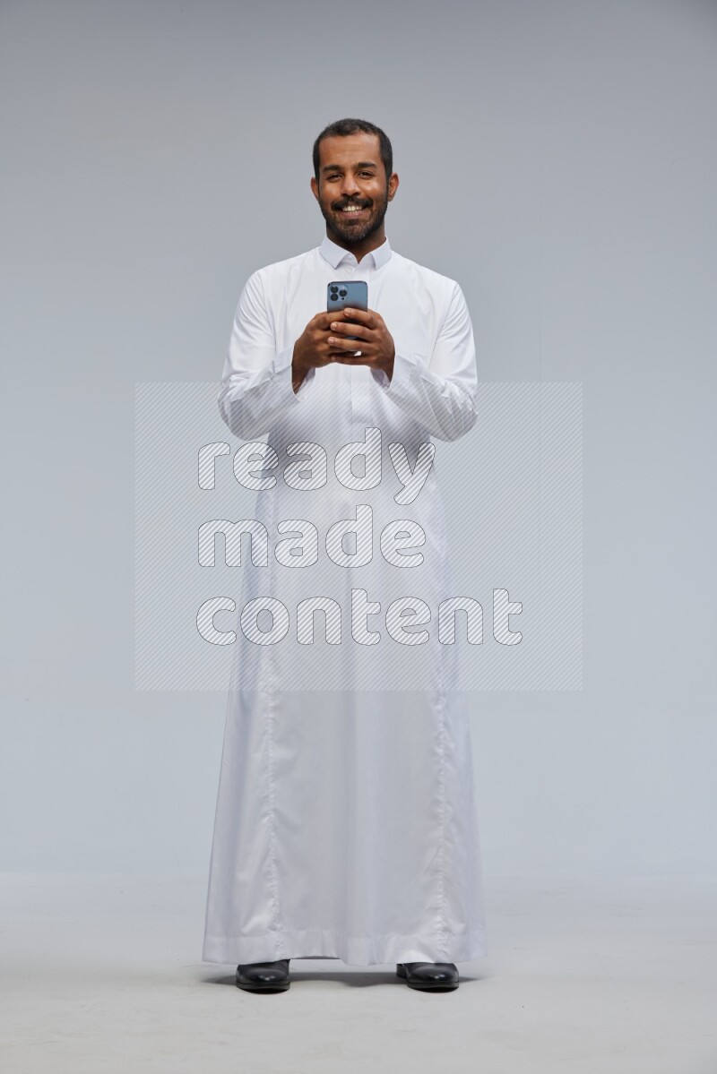 Saudi man wearing Thob standing texting on phone on Gray background