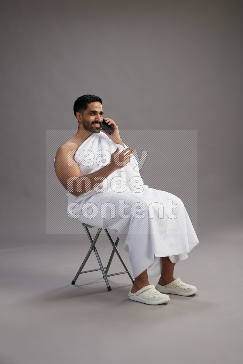 A man wearing Ehram sitting on chair talking on the phone on gray background