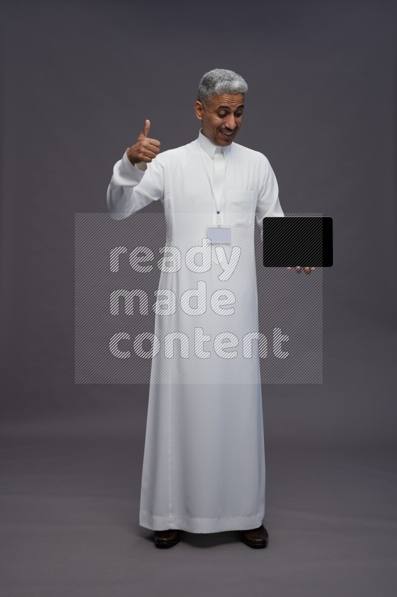 Saudi man wearing thob with neck strap employee badge standing showing tablet to camera on gray background