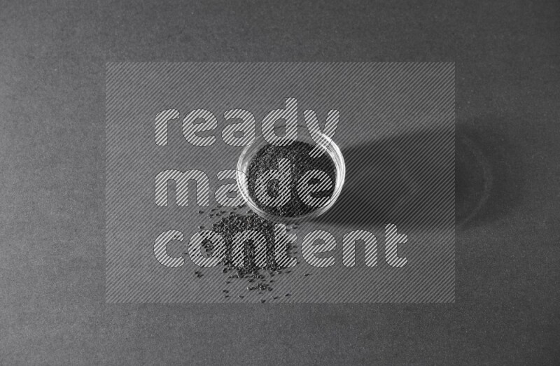 A glass bowl full of black seeds and seeds spreaded beside it on a black flooring