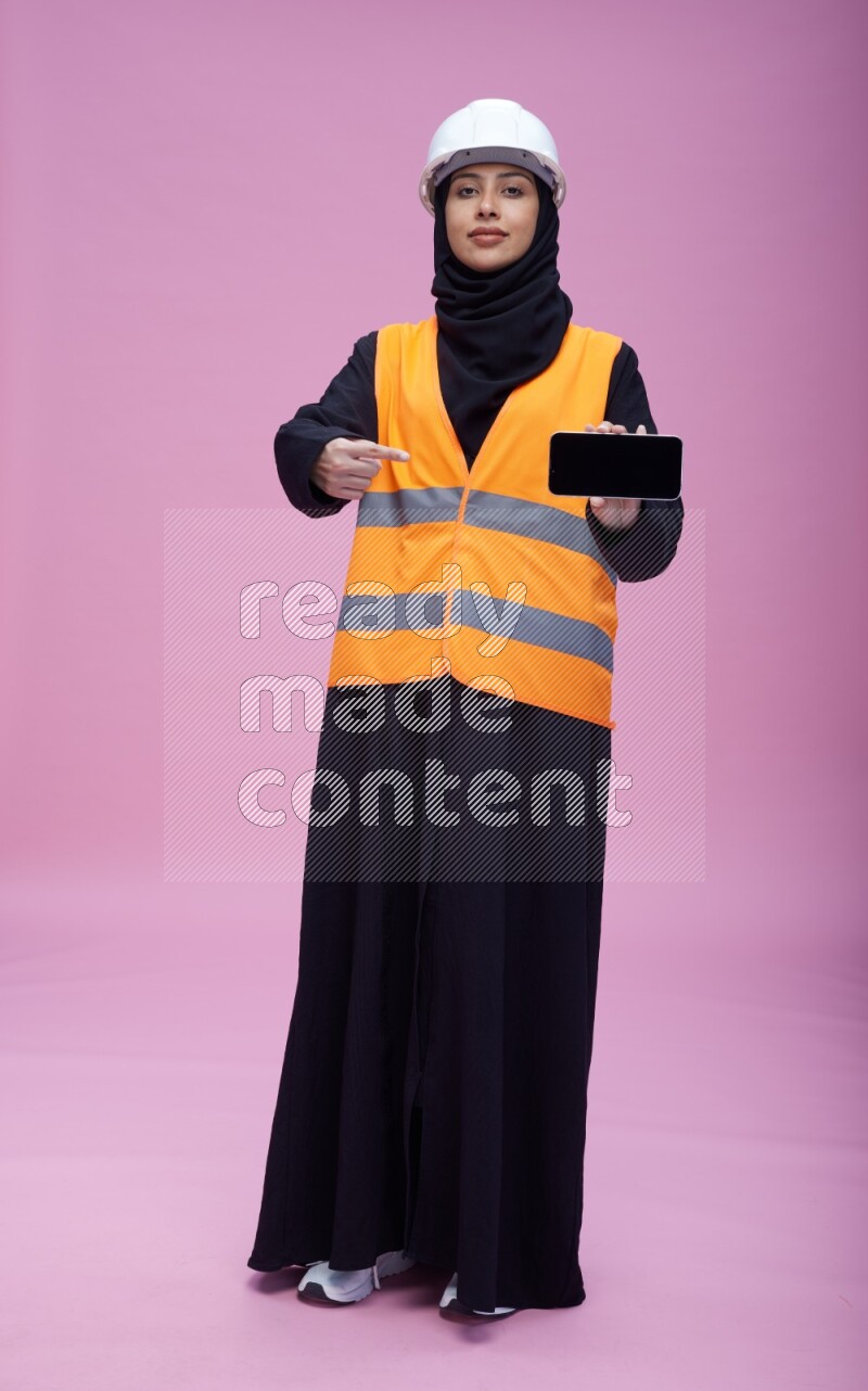 Saudi woman wearing Abaya with engineer vest and helmet standing showing phone to camera on pink background