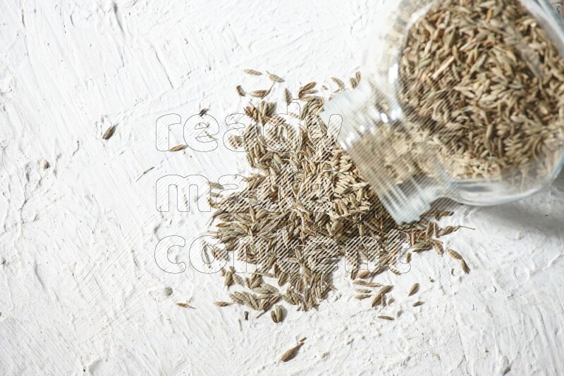 A flipped glass spice jar full of cumin seeds and the seeds spilled out on textured white flooring