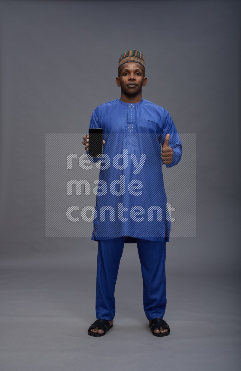 Man wearing Nigerian outfit standing showing phone to camera on gray background
