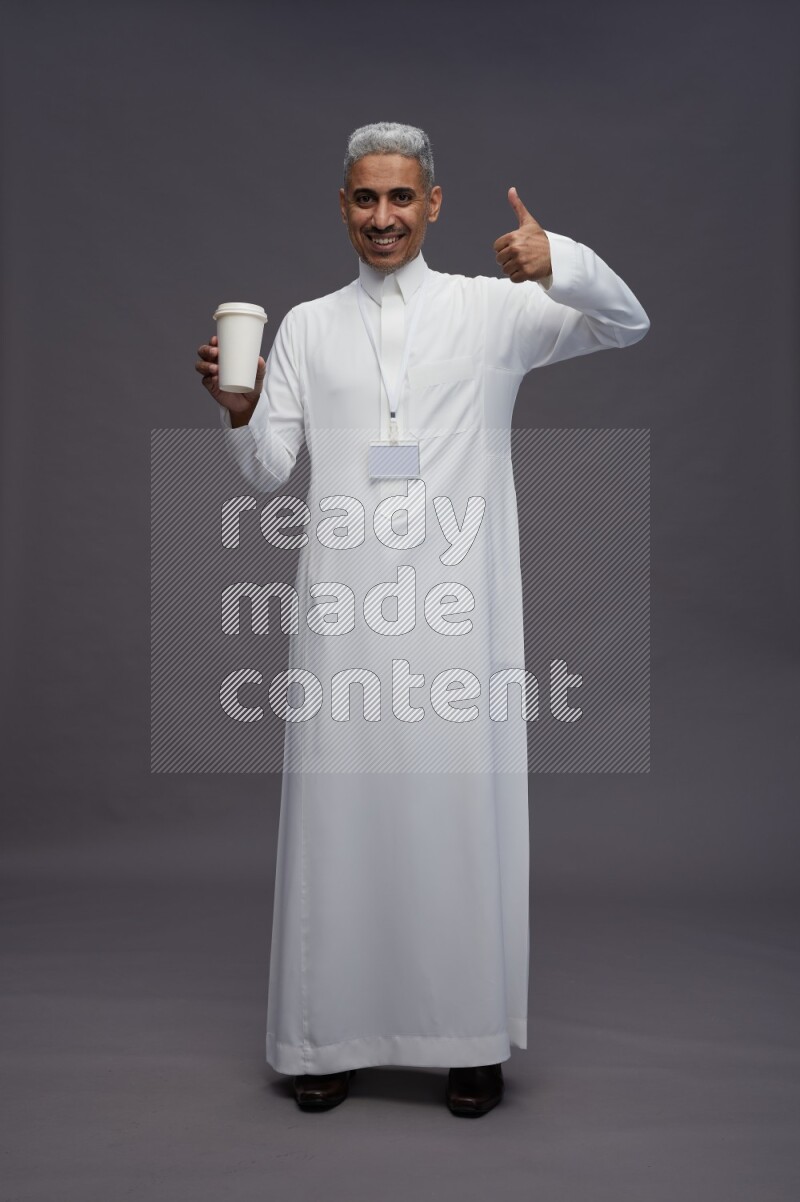 Saudi man wearing thob with neck strap employee badge standing holding paper cup on gray background