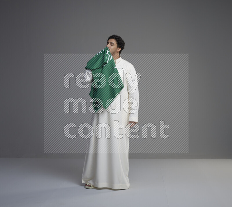 A Saudi man standing wearing thob kissing Saudi flag on gray background