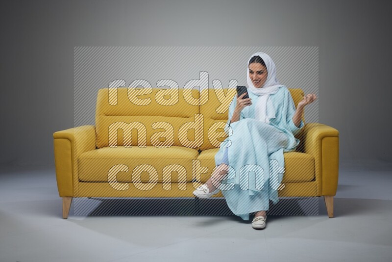 A Saudi woman wearing a light blue Abaya and white head scarf sitting on a yellow sofa and texting on her phone on a grey background