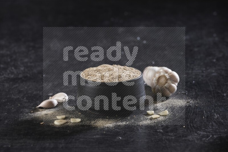 A black pottery bowl full of garlic powder with garlic cloves and a whole bulb beside it on a textured black flooring