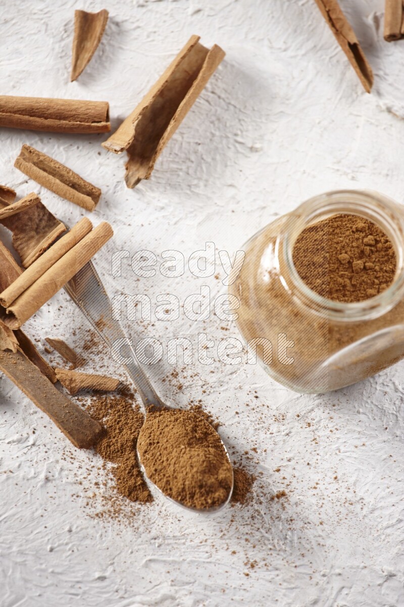 Herbal glass jar full cinnamon powder and a metal spoon surrounded by cinnamon sticks on a white background
