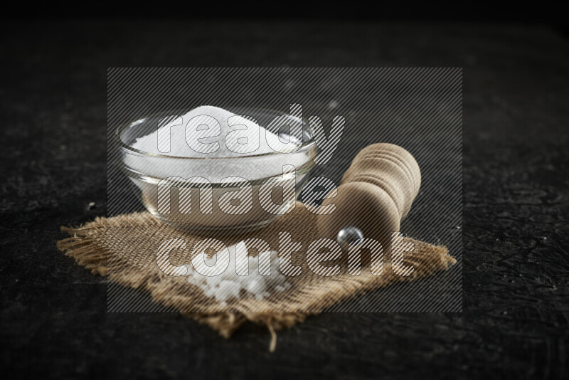 A glass bowl full of white salt with a wooden grinder on a burlap fabric all on black background