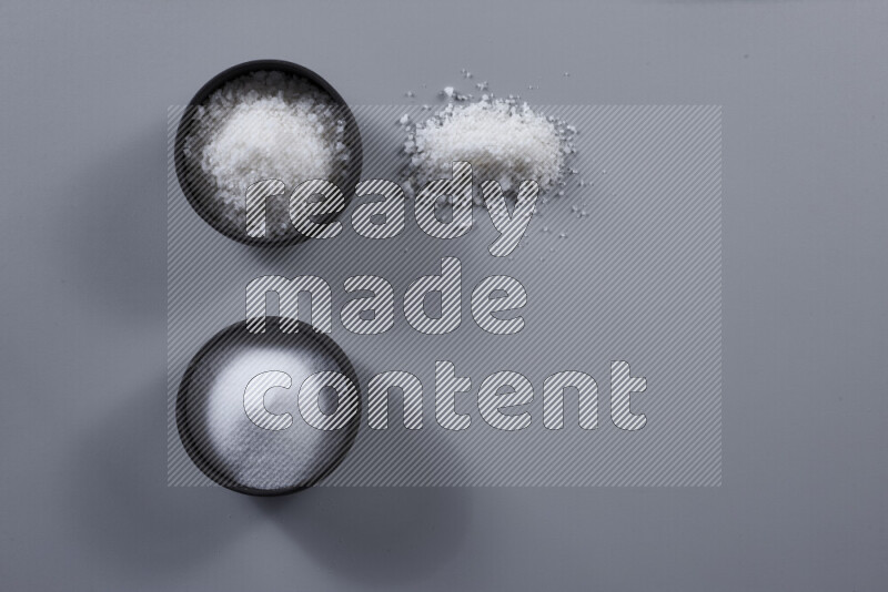 2 pottery bowls one is filled with table salt and the other with coarse sea salt on grey background