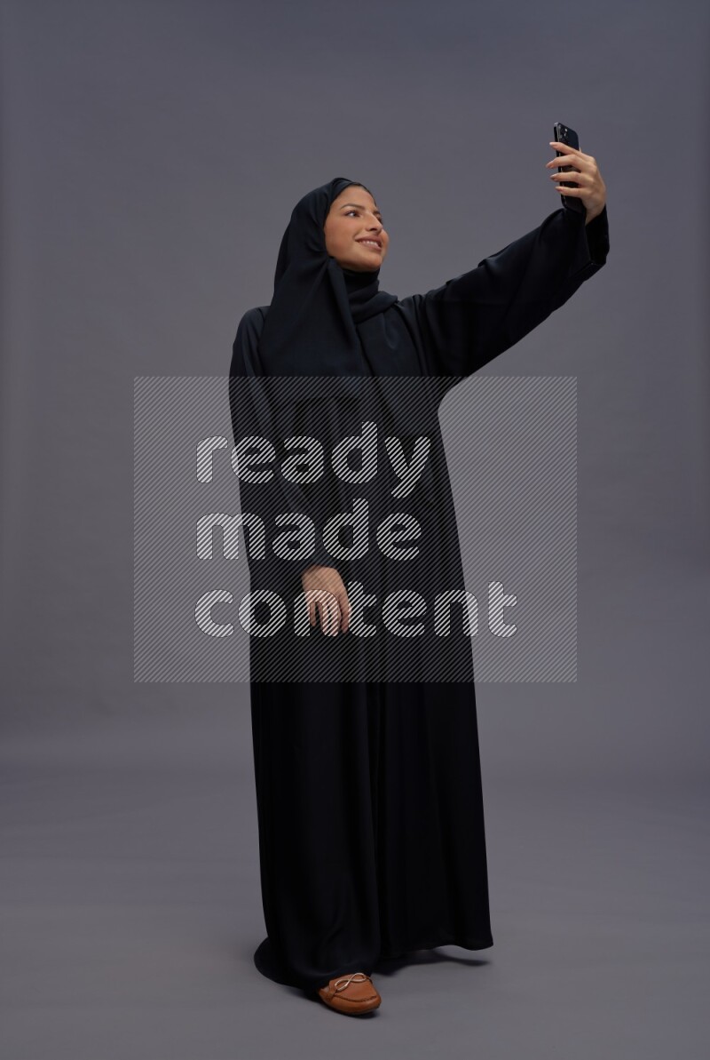 Saudi woman wearing Abaya standing taking selfie on gray background