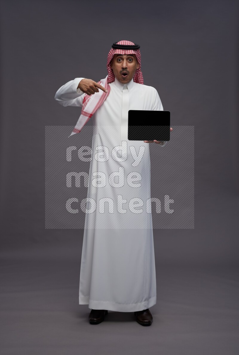 Saudi man wearing thob and shomag standing showing tablet to camera on gray background