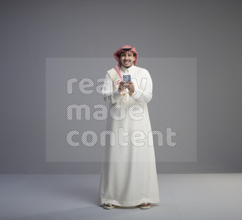 A  Saudi man standing wearing thob and red shomag texting on phone on gray background