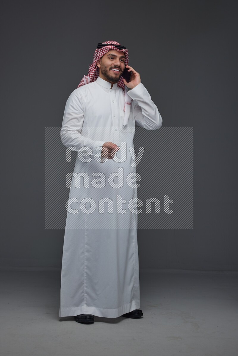 A Saudi man wearing Thobe and Shmagh talking in the phone eye level on a gray background
