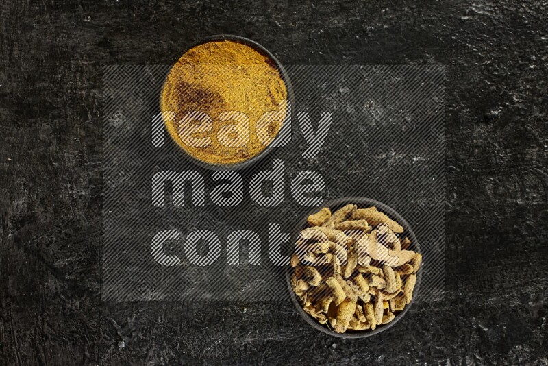 2 pottery black bowls, one full of turmeric powder and the other full of dried turmeric whole fingers on a textured black flooring