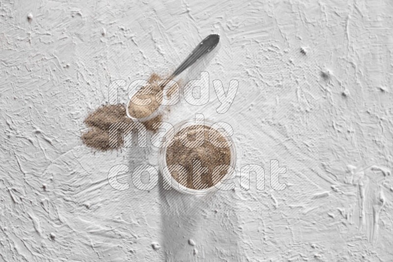 A glass jar full of black pepper powder and a metal spoon full of powder on a textured white flooring