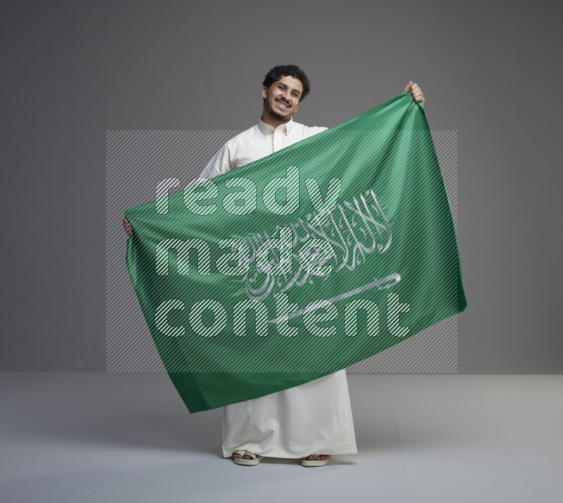 A Saudi man standing wearing thob holding big Saudi flag on gray background