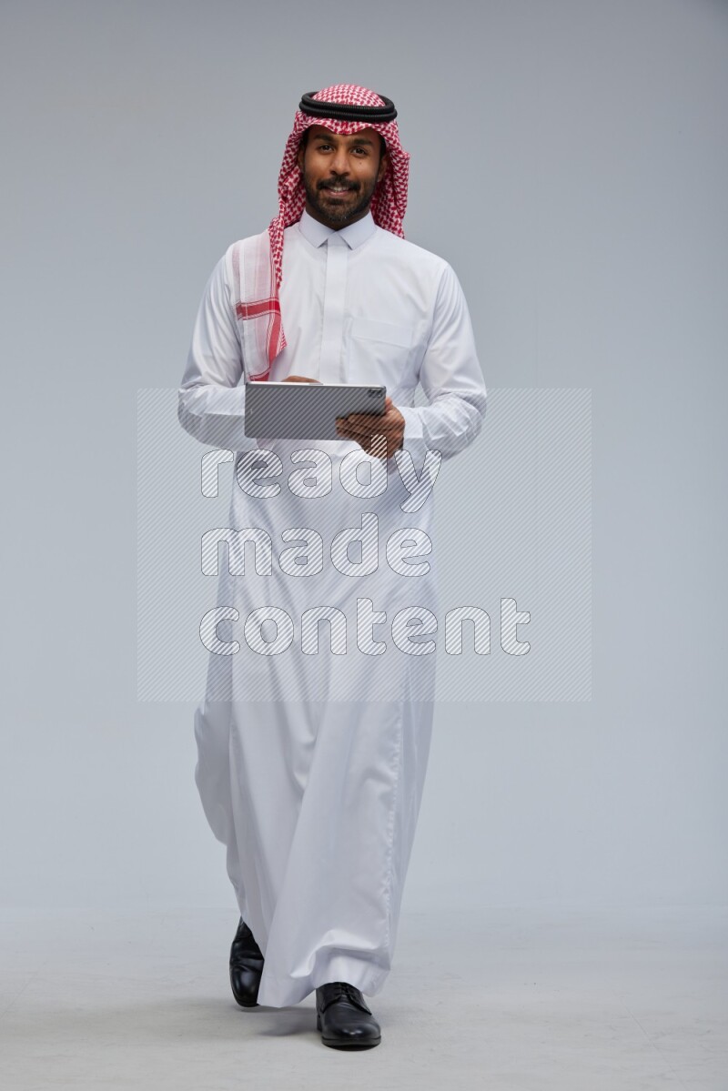Saudi man Wearing Thob and shomag standing working on tablet on Gray background