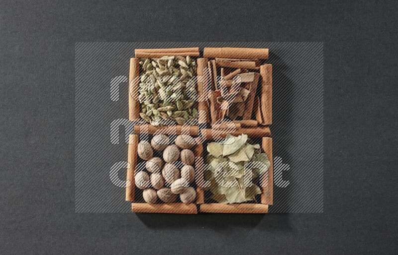 4 مربعات من عيدان القرفة ممتلئة بقطع القرفة وحبوب الهيل وأوراق الغار وجوزة الطيب على أرضية سوداء