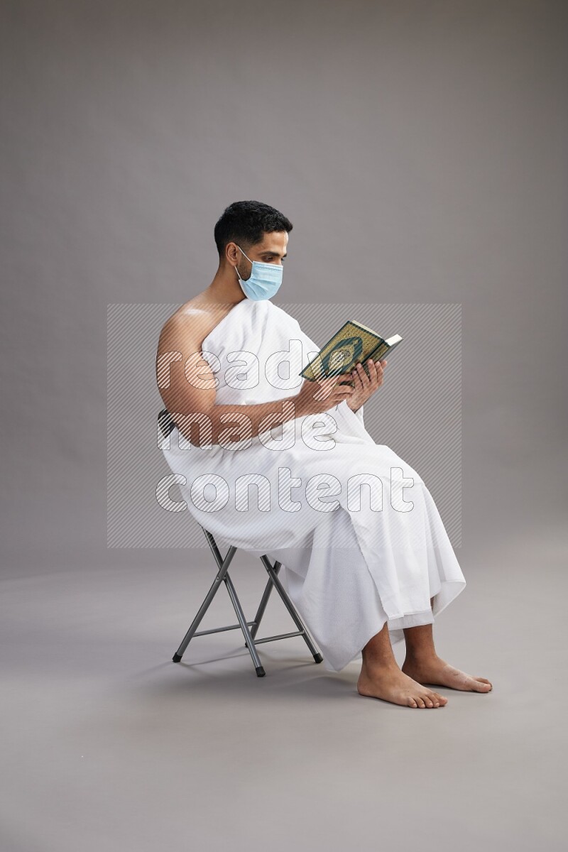 A man wearing Ehram with face mask sitting on chair reading quran on gray background
