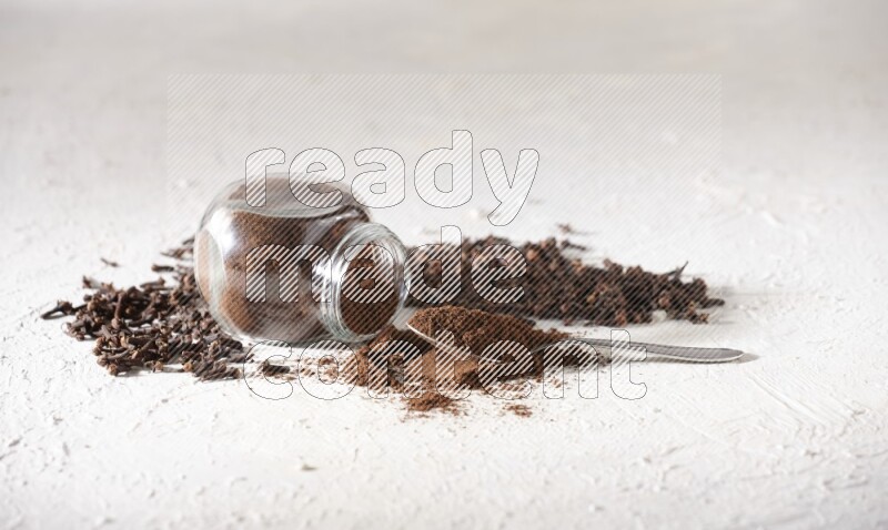 A flipped glass spice jar and a metal spoon full of cloves powder and powder came out of the jar with cloves spread on textured white flooring