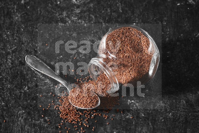 A glass spice jar full of garden cress seeds flipped and seeds spread out with a metal spoon full of the seeds on a textured black flooring