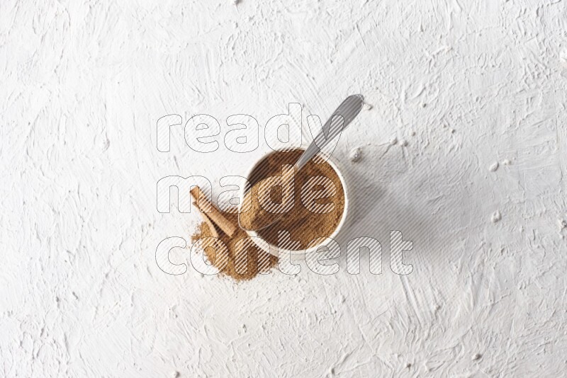 Ceramic beige bowl full of cinnamon powder and a metal spoon with cinnamon sticks next of it on a textured white background
