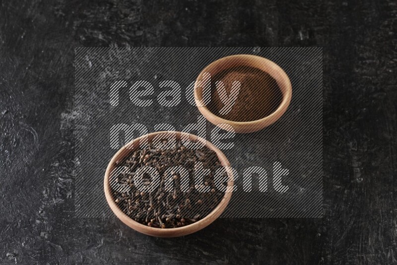2 wooden bowls full of cloves powder and whole cloves on a textured black flooring