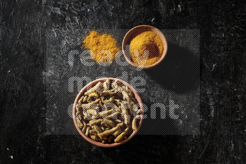 2 wooden bowls, one full of turmeric powder and the other full of dried turmeric whole fingers on a textured black flooring