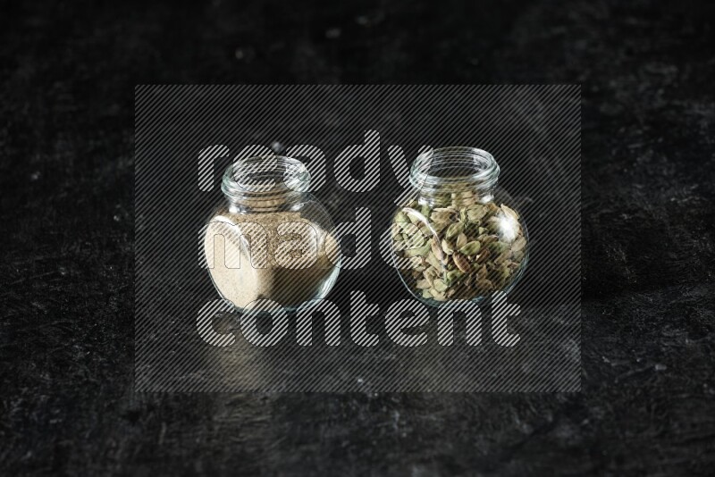 2 glass spice jars full of cardamom powder and cardamom seeds on textured black flooring