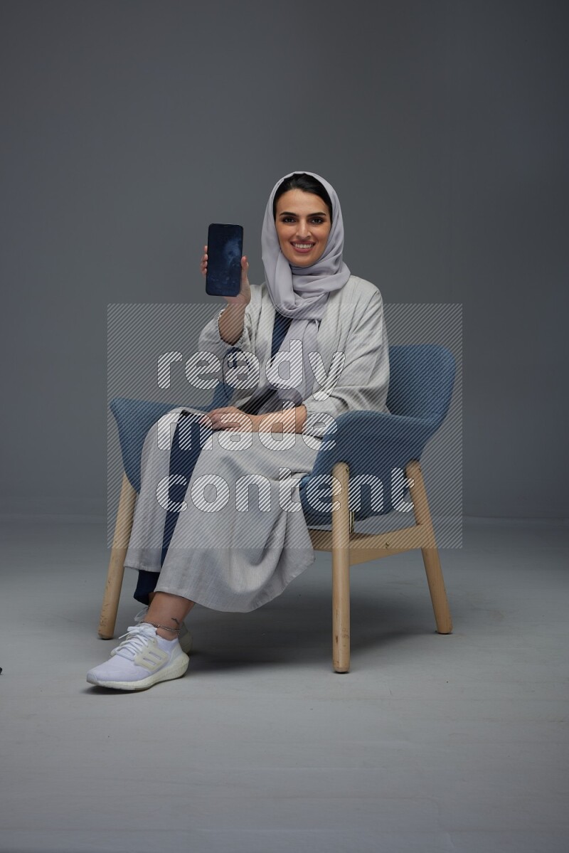 A Saudi woman wearing a light gray Abaya and head scarf sitting on a dark grey chair and showing the phone's screen eye level on a grey background