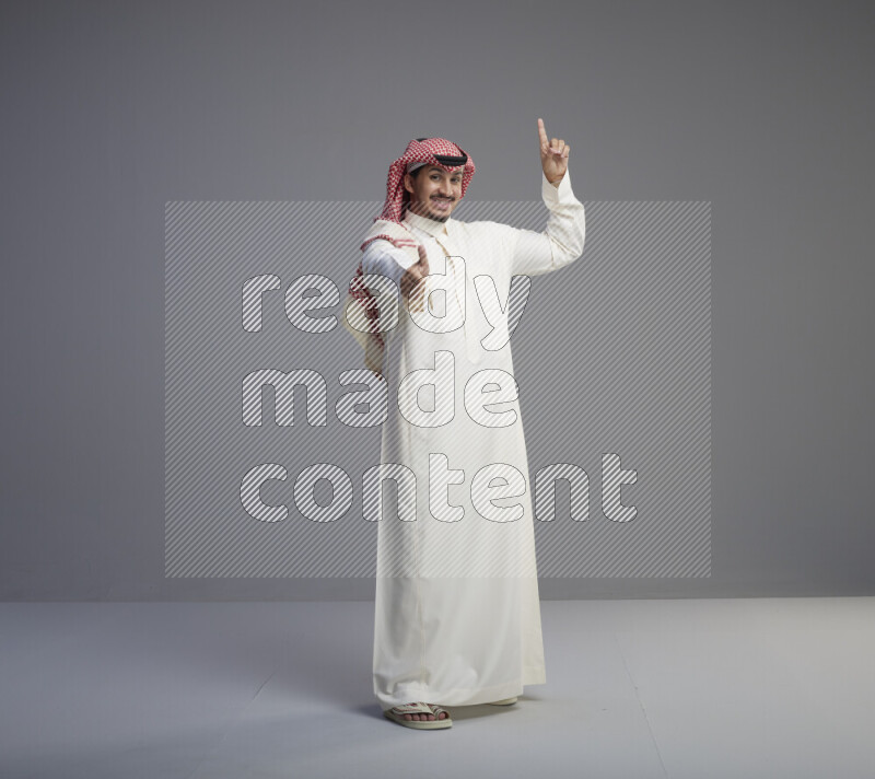 A Saudi man standing wearing thob and red shomag interacting with the camera on gray background