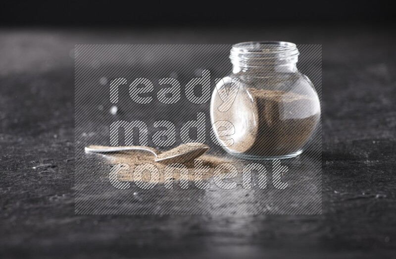 A glass spice jar full of black pepper powder and a metal spoon full of powder on textured black flooring
