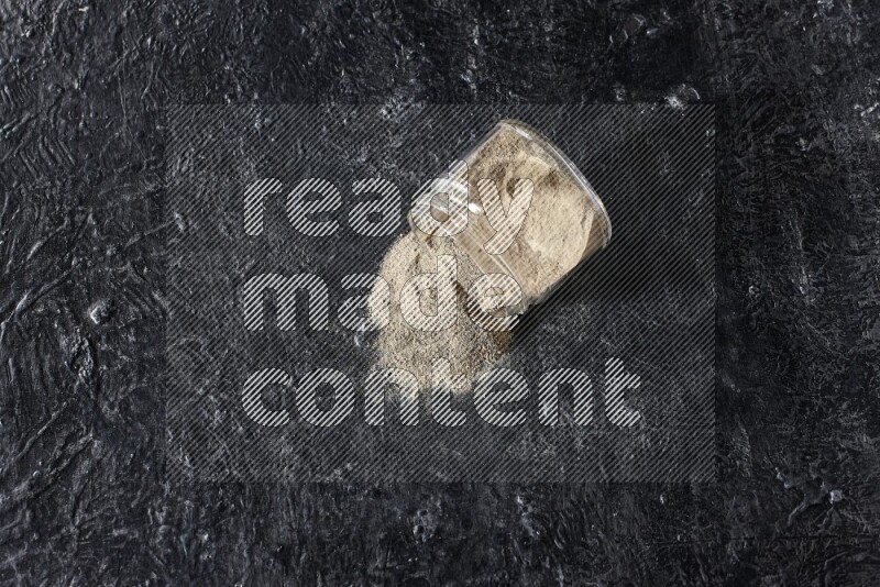 A flipped glass jar full of white pepper powder with spilled powder on textured black flooring