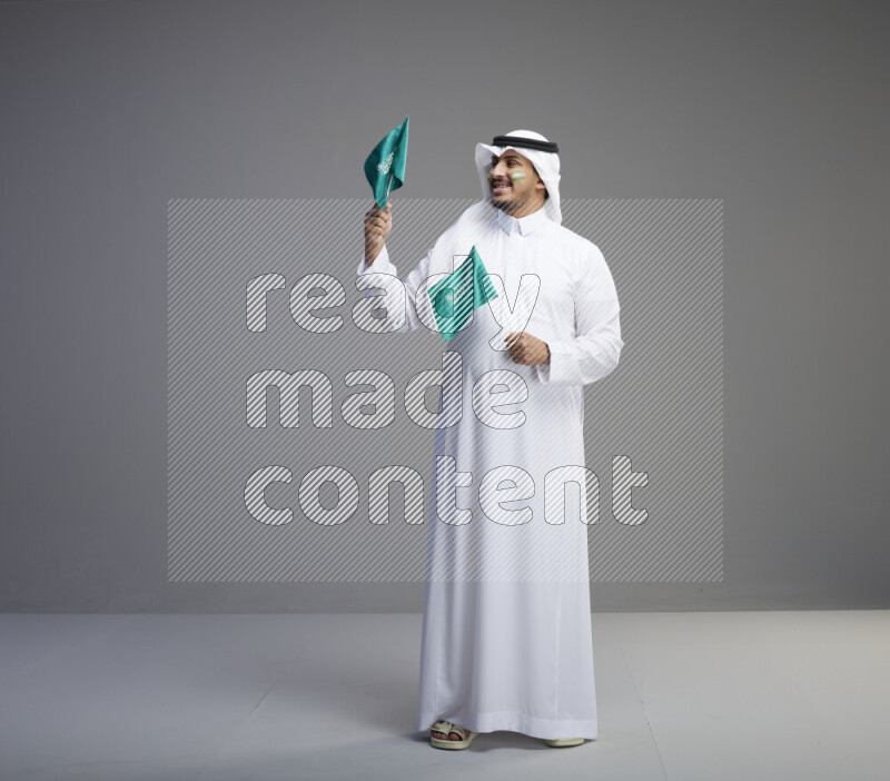 A Saudi man standing wearing thob and white shomag with face painting raising small saudi flag on gray background