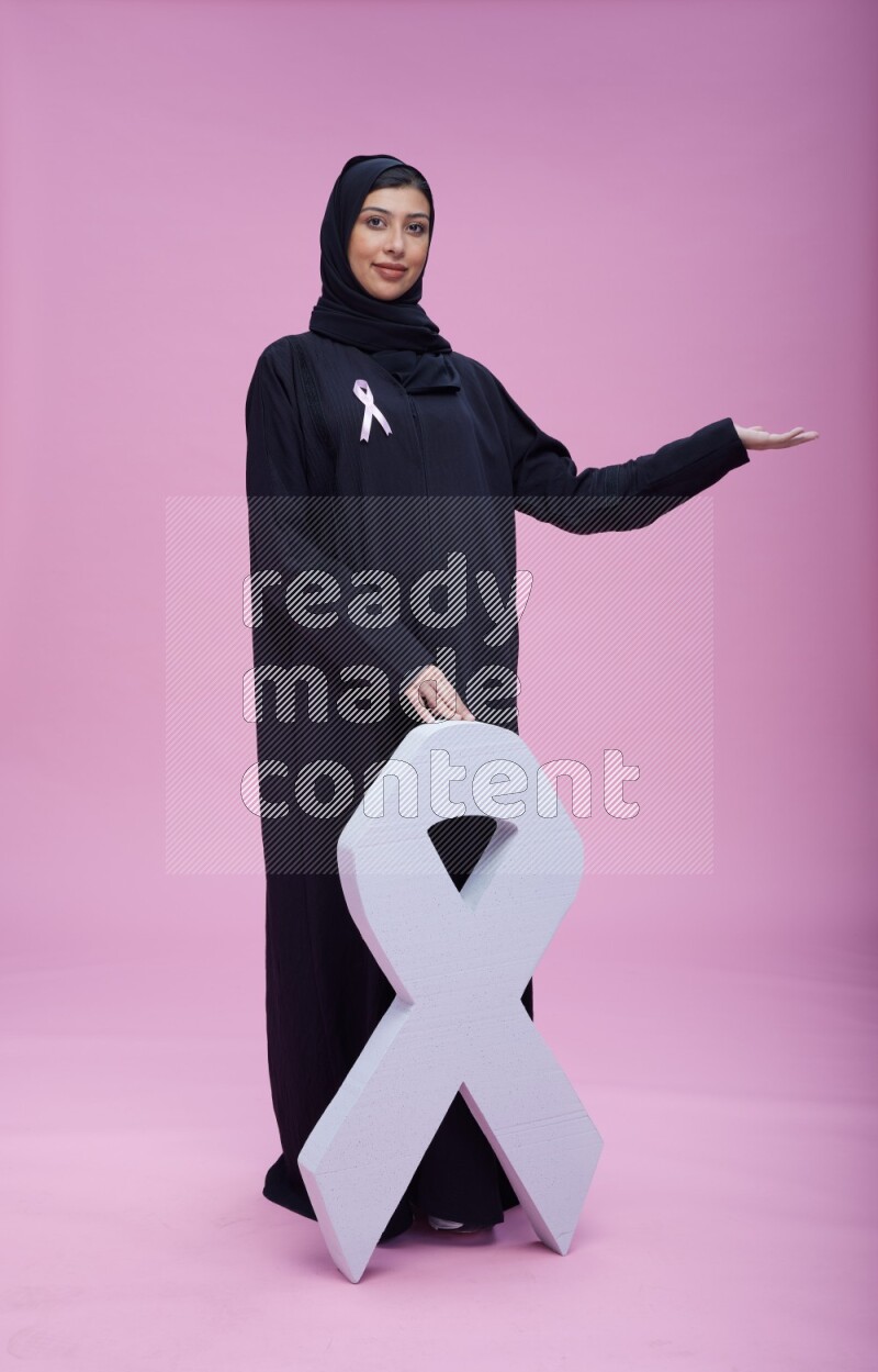 Saudi woman wearing pink ribbon on abaya standing holding awareness ribbon on plain pink background