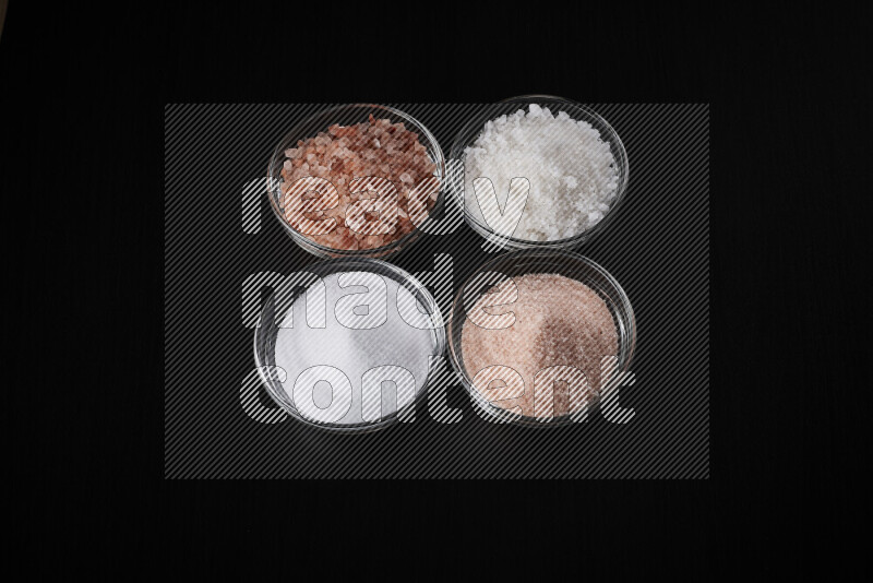 4 glass bowls filled with table salt, coarse sea salt, fine himalayan salt and coarse himalayan salt on black background